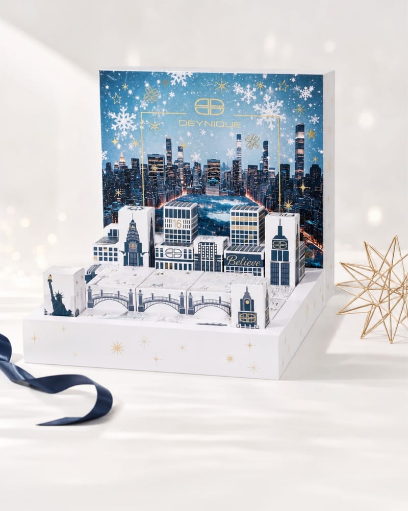 Der aufgeklappte DEYNIQUE Adventskalender 2026 mit Skyline-Rückwand, Türchen-Häusern und goldenem Stern